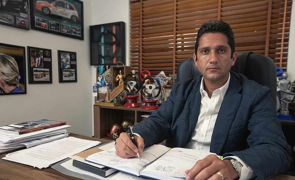 Douglas Carvalho Jr. na mesa do escritório, com fotos de corrida e troféus atrás